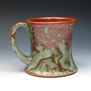 May include: A handmade ceramic mug with a unique design. The mug features a red interior and rim, with a mottled red and green exterior. The handle is green with red accents. The base is a natural, unglazed color. A functional and decorative piece.