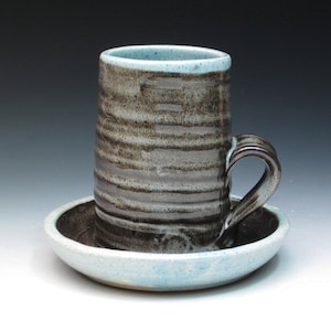 May include: A ceramic mug with a blue and brown glaze and a matching saucer. The mug has a handle and is decorated with a striped pattern.
