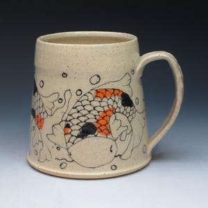 May include: A beige ceramic mug with a large handle, featuring a hand-drawn design of koi fish. The fish have orange and black scales, with black outlines. Bubbles and aquatic plants are also drawn around the fish.