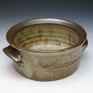 May include: A handmade ceramic bowl with two handles. The bowl has a wide rim with a reddish-brown edge and a light brown interior. The exterior is a mottled gray-brown with subtle variations in color, giving it a rustic appearance.