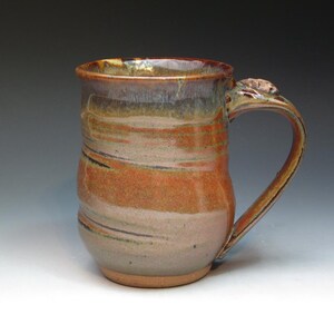May include: A handmade ceramic mug with a brown, orange, and grey glaze. The mug has a rounded body and a handle with a small decorative detail.