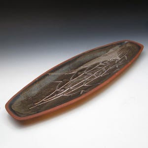 May include: A long, handmade ceramic serving dish with a dark brown interior and a reddish-brown rim. The dish features an etched bamboo design. The elongated shape is ideal for serving appetizers or displaying decorative items.