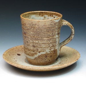 May include: A brown and white ceramic coffee cup and saucer with a speckled glaze. The cup has a rounded shape and a handle. The saucer is round and has a slightly raised edge.