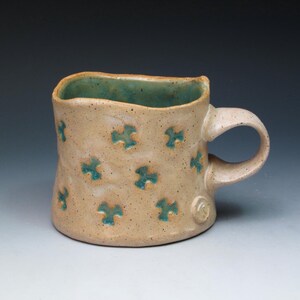 May include: Handmade ceramic mug with a tan exterior and a teal interior. The mug features a series of teal-colored, four-petal flower designs. The handle is curved and the mug has a unique, textured surface. A perfect addition to any kitchen.