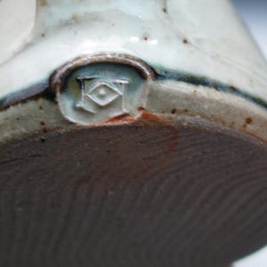 May include: Close-up of the bottom of a ceramic pot with a brown glaze. The pot has a maker's mark in the center of the bottom, which is a circle with the letters "K" and "H" inside, and a small circle in the center.