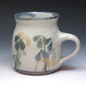 May include: Handmade ceramic mug with a cream-colored base and a blue rim and base. The mug features a floral design in shades of blue and yellow. The mug has a curved handle.