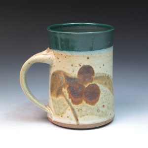 May include: A ceramic mug with a green glaze on the top and a brown and white glaze on the bottom. The mug has a handle and a brown design of berries on the side.