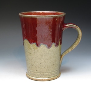May include: A ceramic mug with a red glaze on the top half and a speckled beige glaze on the bottom half. The mug has a rounded handle.