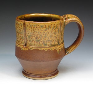 May include: A handmade ceramic mug with a textured, mottled surface in shades of brown, tan, and green. The mug features a sturdy handle and a wide, cylindrical body, perfect for enjoying hot beverages.