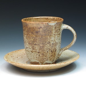 May include: A speckled brown and cream ceramic coffee mug with a handle, sitting on a matching saucer.