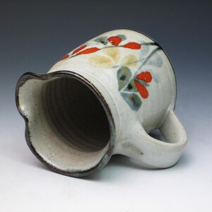 May include: A white ceramic pitcher with a brown rim and a floral design featuring red, yellow, and green accents. The pitcher has a handle and a wide opening.