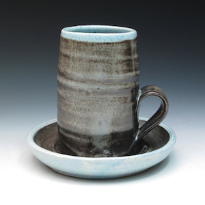May include: A handmade ceramic mug and saucer set. The mug is cylindrical with a dark handle and a light blue rim. The exterior has a textured, layered appearance in shades of gray and brown. The saucer is a matching light blue with a dark interior.