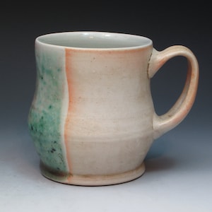 May include: A ceramic mug with a green and white glaze on one side and a light brown glaze on the other side. The mug has a rounded body and a handle.