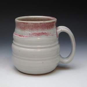 May include: A white ceramic mug with a pink and white glaze. The mug has a rounded body and a handle. The mug is handmade and has a rustic look.