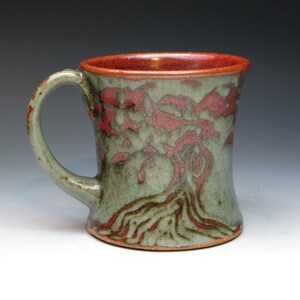 May include: Handmade ceramic mug with a tree design. The mug features a green exterior with a red tree pattern and a red interior. The handle is curved and the mug has a wide base. A unique piece for coffee or tea.