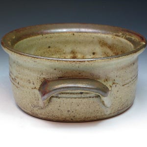 May include: A handmade ceramic bowl with a speckled, light beige exterior and a darker brown rim. The bowl features two small, integrated handles and is suitable for serving or display. The rustic design adds a touch of natural elegance.