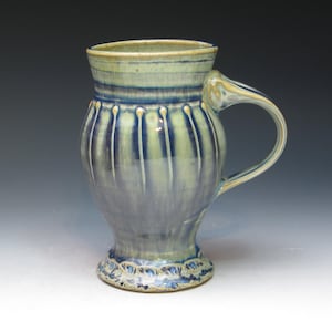 May include: A handcrafted ceramic mug with a distinctive design. The mug has a blue and green glaze, vertical grooves, and a decorative base. The handle is curved, and the rim is slightly flared. A lovely piece of pottery for enjoying a hot drink.