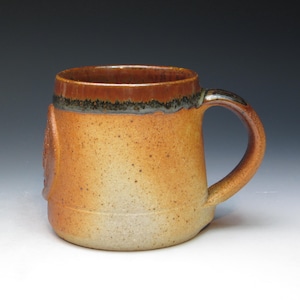 May include: A handmade ceramic mug with a speckled, earthy color palette. The mug features a wide, cylindrical body with a rounded handle. The rim is a darker brown, and the body transitions from a light beige to a warm, speckled orange. The mug has a rustic, artisanal feel.