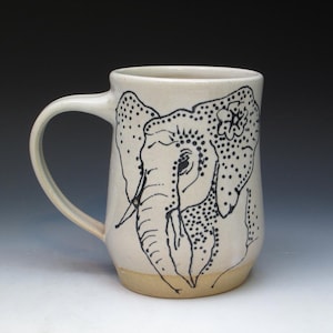 May include: Cream-colored ceramic mug featuring a black elephant illustration. The elephant's head is depicted with dotted details and outlined features. The mug has a curved handle and a light brown base.
