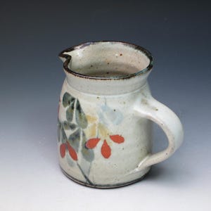 May include: A small, white ceramic pitcher with a brown rim and handle. The pitcher is decorated with a floral design in shades of green, red, and yellow.