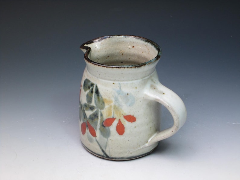 May include: A small, white ceramic pitcher with a brown rim and handle. The pitcher is decorated with a floral design in shades of green, red, and yellow.
