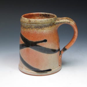 May include: A handmade ceramic mug with a large handle. The mug features a gradient of colors, transitioning from a light beige at the base to a reddish-orange in the middle and a brown rim. Black, abstract lines wrap around the mug.