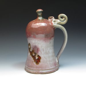 May include: A hand-crafted ceramic salt shaker with a pink and white glaze. The shaker has a unique handle with a curled top and a small hole in the lid for dispensing salt.