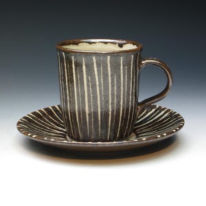 May include: A ceramic coffee cup and saucer set. The cup and saucer are dark brown with vertical white stripes. The cup has a handle and the saucer is round. The interior of the cup is off-white.