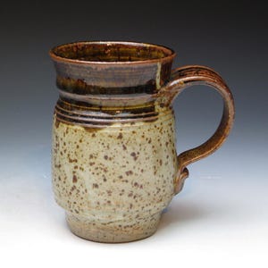 May include: A brown and white speckled ceramic mug with a brown handle. The mug has a wide rim and a rounded body.