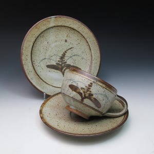 May include: A handmade ceramic teacup, saucer, and plate set. The set features a speckled beige glaze with brown and blue botanical designs. The cup is tilted on its saucer, with the plate in the background.