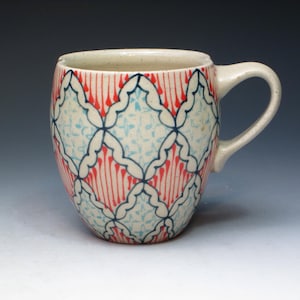 May include: A white ceramic mug with a red and blue geometric pattern. The pattern features a diamond shape with a floral design inside.