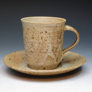 May include: A speckled beige ceramic coffee mug with a handle, sitting on a matching saucer.