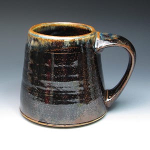 May include: A handmade ceramic mug with a dark, glossy finish. The mug has a wide base that tapers upwards, with a large handle. The rim has a brown and blue glaze. The mug is approximately 13 cm tall.