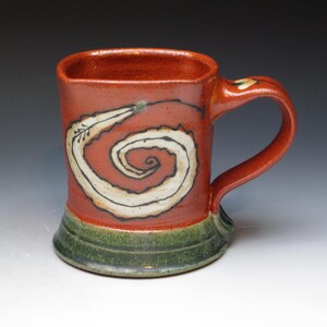 May include: A handmade ceramic mug with a square top and a red-orange glaze. A white spiral design with black outlines decorates the front. The base is green, and the handle is red-orange. This unique mug is perfect for coffee or tea.