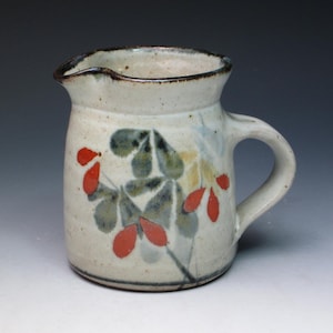 May include: A small white ceramic pitcher with a brown rim and a handle. The pitcher is decorated with a floral design in shades of green, red, and yellow.