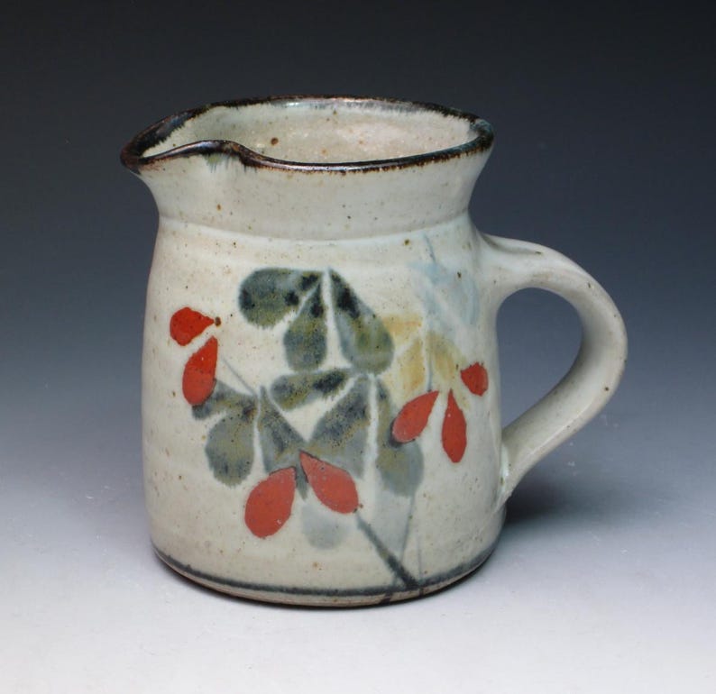 May include: A small white ceramic pitcher with a brown rim and a handle. The pitcher is decorated with a floral design in shades of green, red, and yellow.