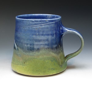 May include: Handmade ceramic mug with a blue and green glaze. The mug features a rounded handle and a wide, slightly flared body. The colours blend from a deep blue at the top to a vibrant green at the base, creating a gradient effect. A unique piece for enjoying beverages.