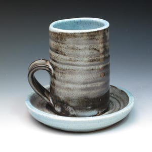 May include: A ceramic mug with a dark brown and gray glaze, with a light blue interior and a matching saucer. The mug has a rounded handle.
