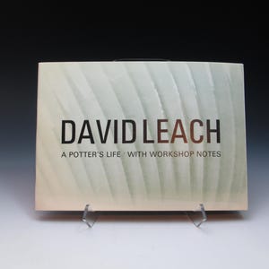 May include: A book titled "DAVID LEACH" with the subtitle "A Potter's Life with Workshop Notes." The cover has a light green and white textured design. The book is displayed on a clear stand against a dark background.