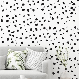 May include: A living room interior with a white sofa and two cushions, a white coffee table, a small wooden side table with a vase of greenery, and a white wall with a black and white dalmatian spot pattern.