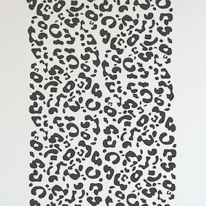 Op de afbeelding: Een witte muur met een groot, verticaal patroon van donkergrijze luipaardvlekken. Een kleine witte tafel, stoel en decoratieve items staan op de voorgrond. Een knuffel pinguïn zit op de stoel.