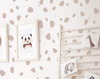 May include: A white crib with a white blanket and pillows in a nursery with a white wall featuring a brown pebble pattern. There is a white stool, a white ladder shelf with pictures, and a wooden stool with the word "baby" on it.