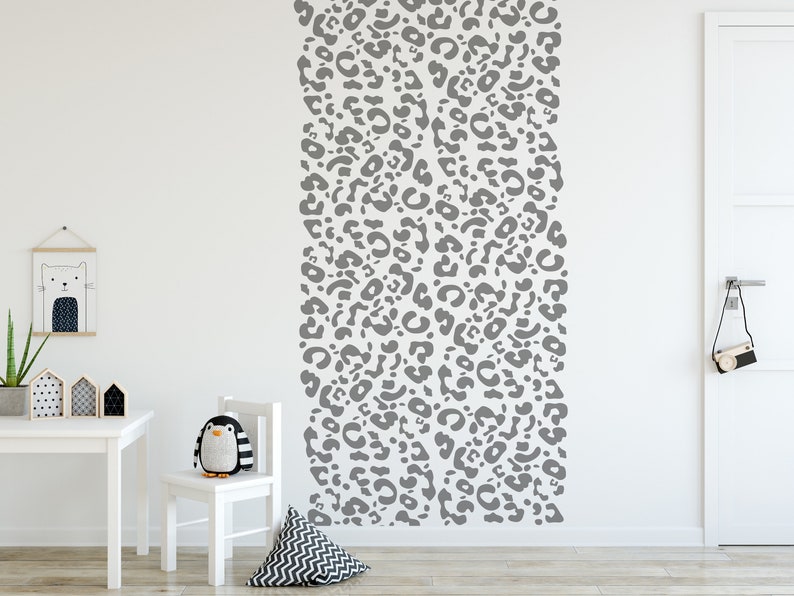 May include: A white wall with a vertical gray leopard print design. A white table, chair, and decorative items are in the foreground. A stuffed penguin sits on the chair. A framed cat illustration hangs on the wall.