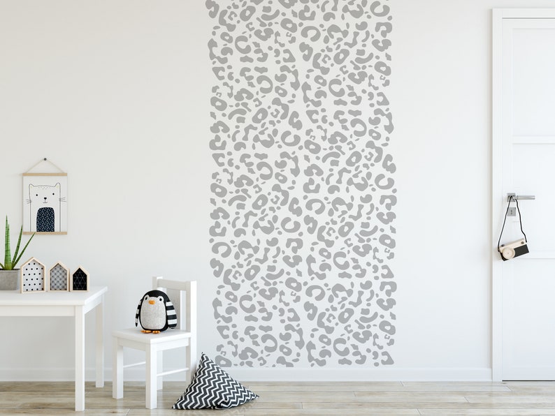 May include: A white wall with a vertical pattern of gray leopard print. A white table, chair, and decorative items are in the foreground. A stuffed penguin sits on the chair. A framed cat illustration hangs on the wall.