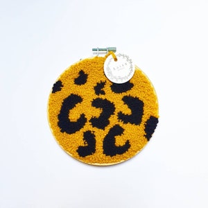Può includere: Un tappeto rotondo in lana giallo e nero, cucito a mano, con un motivo a stampa leopardata. Il tappeto è montato su un cerchio di legno e ha un piccolo cartellino bianco con il testo "BAIRN" su di esso.
