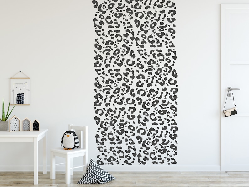 May include: A white wall with a large, vertical pattern of dark gray leopard spots. A small white table, chair, and decorative items are in the foreground. A stuffed penguin sits on the chair.