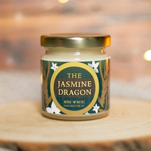 May include: A small glass jar candle with a gold lid. The label is green with gold accents and features a white floral design. The text on the label reads "The Jasmine Dragon" and "Nerdie Nifties Vegan Cruelty Free Soy".
