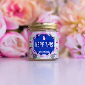 May include: A clear glass candle jar with a gold lid. The label is pink, green, and blue with a white bunny and the text "NERF THIS", "NERDIE", "NIFETIES", "VEGAN", "CRUELTY FREE SOY", and "120g".