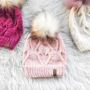 May include: Three knitted hats with faux fur pom poms. The hats are pink, white, and a deep pink with a purple hue. The hats have a cable knit pattern and a small leather tag with the text "A Knit Life" on them.