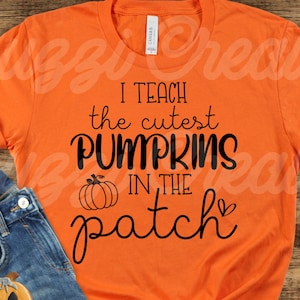May include: Orange t-shirt with black text that reads "I teach the cutest pumpkins in the patch". The shirt is laying on a wooden surface with a pair of blue jeans, a ghost, and a pumpkin.